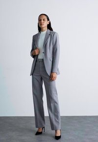 Fato cinza ajustado com um blazer entallado e calças de perna larga, combinado com uma blusa de tom claro e saltos altos pretos. Textura de tecido suave.