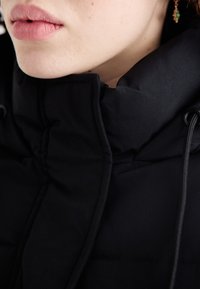 Black padded jacket with a high collar, featuring drawstrings and a smooth, matte texture. Close-up of lips and skin tone.