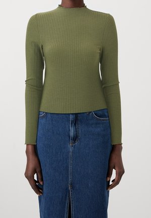 Personne portant un haut côtelé à manches longues vert olive et une jupe en jean bleue avec une fente à l'avant, debout devant un fond uni.