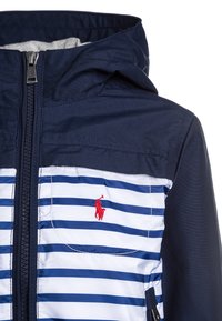 Námořnická modrá bunda s vodorovnými bílými a modrými pruhy, zipem vpředu, kapucí a malým červeným logem na kapsičce na prsou.