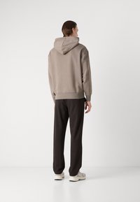 Sudadera marrón claro con una costura trasera y capucha ajustable, combinada con pantalones negros y zapatos claros. Diseño sencillo, textura suave.
