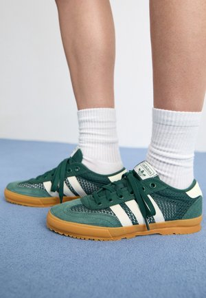 Person iført hvide ribstrikkede sokker og grønne Adidas-sneakers med hvide striber og gummisåler, stående på et lyseblåt tæppe.