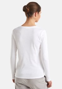 Kobieta z brązowymi włosami w kok w białej, gładkiej bluzce z długim rękawem i jasnych spodniach, odsunięta plecami na białym tle.