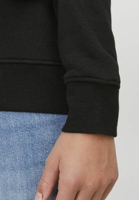 Sudadera negra con puños y dobladillo acanalados. Se muestra con jeans de mezclilla azul claro, resaltando la suave textura y el diseño ajustado.