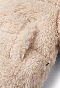 Miękka, kręcona beżowa tkanina polarowa z szwem i częściowo widoczną czarną metką z boku.