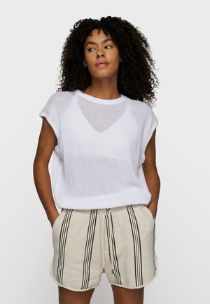 Frau mit lockigem Haar, die ein weißes ärmelloses Netzoberteil und beige Shorts mit vertikalen schwarzen Streifen und Seitentaschen trägt, mit den Händen in den Taschen.