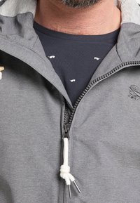 Harmaa vetoketjullinen takki, jossa on huppu ja teksturoitu pinta sekä valkoinen kiristysnauha. Alla tummansininen paita, jossa pieniä valkoisia kuvioita.