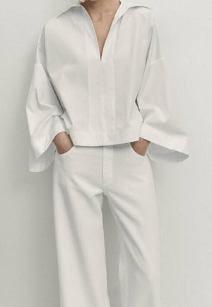 Person iført løs hvid bluse med brede ærmer og vidde bukser, hænderne afslappet i lommerne, stående mod ensfarvet baggrund.