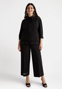 Camicia nera a maniche tre quarti con bottoni e rifiniture decorative sulle spalle, abbinata a pantaloni larghi con dettagli a taglio.