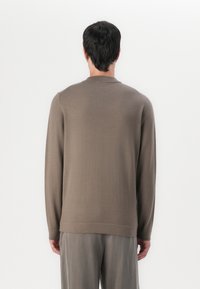 Maglione marrone a maniche lunghe realizzato in morbido tessuto a maglia, con un classico scollo rotondo e una vestibilità rilassata, indossato con pantaloni grigi chiari.