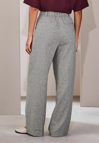 Persona con pantalones grises holgados con cintura elástica y zapatos beige pálido, de pie contra paredes neutrales claras.