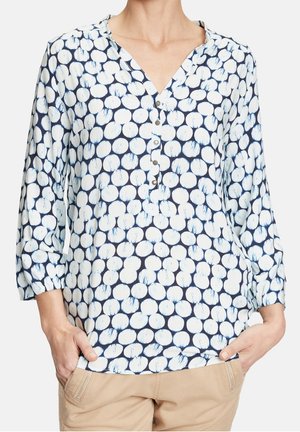 Mujer con blusa azul marino con lunares blancos y mangas tres cuartos y pantalones beige, manos en los bolsillos.