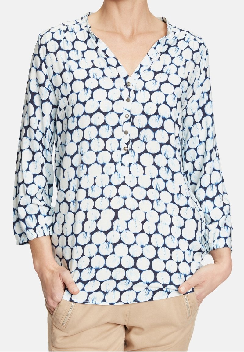 Femme portant un chemisier bleu marine à pois blancs avec des manches trois-quarts et un pantalon beige, mains dans les poches.