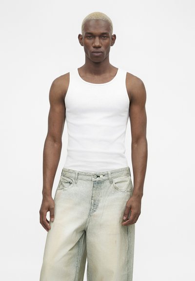 Άνδρας με κοντά ξανθά μαλλιά που έχουν υποστεί λεύκανση, φορώντας λευκό αμάνικο τοπ και ανοιχτόχρωμο, άνετο τζιν, στέκεται μπροστά σε λευκό φόντο.