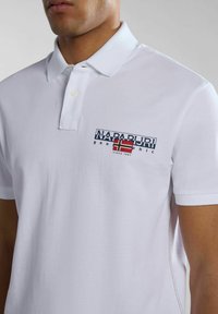 Witte polo met een twee-knopen knoopsluiting, voorzien van een marineblauw en rode logo op de borst. Zacht katoenen stof met textuurdetail.