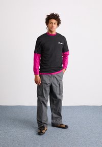 T-shirt noir à manches courtes sur une chemise rose à manches longues, associé à un pantalon cargo gris et des baskets noires avec des accents de léopard.