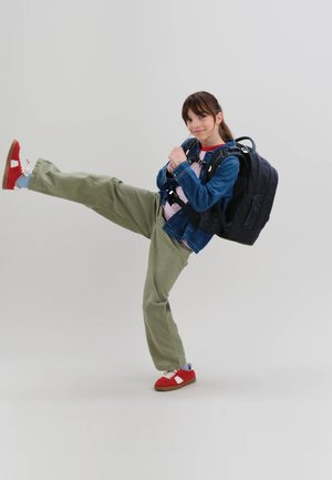 Mädchen trägt grüne Hose, Jeansjacke, rote Turnschuhe und schwarzen Rucksack, hebt ein Bein hoch in einer verspielten Pose vor schlichtem Hintergrund.