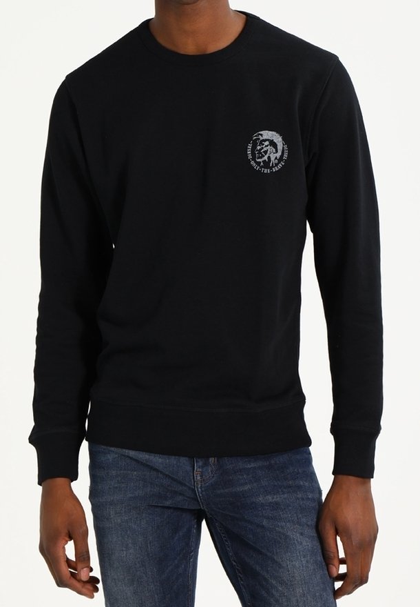 Homme portant un t-shirt noir à manches longues avec un petit logo graphique sur la poitrine gauche et un jean bleu.
