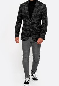 Czarna marynarka w kamuflaż z dwoma guzikami, zestawiona z szarymi dżinsami skinny i czarnymi sneakersami. Marynarka ma klapy w kształcie litery V oraz dwie przednie kieszenie.