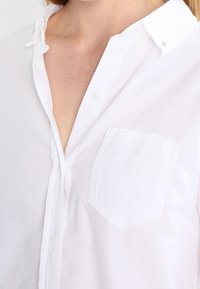 Femme portant une chemise blanche à col déboutonnée avec une poche sur la poitrine, montrant une partie de son cou et de son haut de la poitrine.