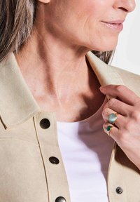 Veste en suède beige avec des boutons noirs, portée sur un haut blanc. Main ornée de deux bagues avec des pierres vertes et aqua.