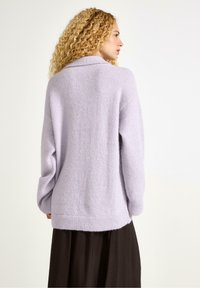 Donna con capelli biondi ricci che indossa un maglione larghi color lavanda e una lunga gonna scura, rivolta di lato con la schiena per lo più visibile.