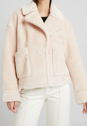 Vrouw draagt een lichtroze teddywollen jack met een grote kraag en opgestikte zakken aan de voorkant, gecombineerd met een witte broek en een zwarte top.