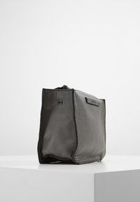 Bolso de cuero gris con acabado texturizado, forma rectangular y cremallera superior. Presenta una marca minimalista y un diseño liso y sin adornos.
