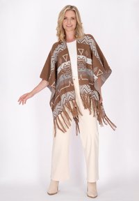 Brauner Strick-Poncho mit geometrischen Mustern in Creme und Grau, mit Fransen an den Kanten. Getragen über einem weißen Oberteil und beigen Hosen.