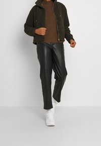 Braune, fuzzy Zip-Jacke über einem strukturierten braunen Pullover, kombiniert mit schwarzen Lederhosen und weißen Sneakers. Schlichtes, modernes Design.