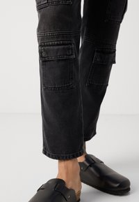 Pantaloni cargo neri a gamba dritta, con molteplici tasche con pattina, realizzati in denim. Indossati con zoccoli neri a slip-on con suola in sughero.