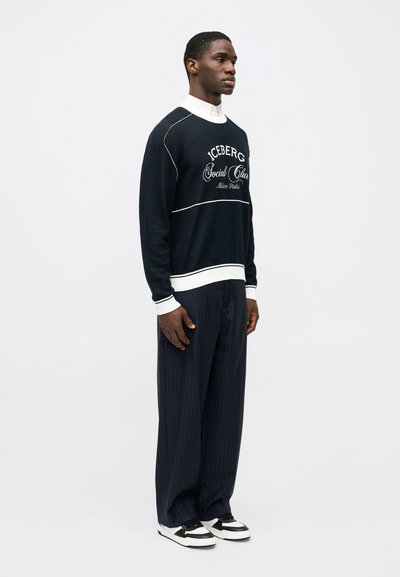 Άνδρας που φοράει μαύρη και άσπρη μπλούζα με το κείμενο "Iceberg Social Club", φαρδύ παντελόνι με ρίγες, λευκή πουκάμισο και μαύρα-άσπρα αθλητικά παπούτσια.