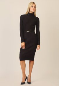 Schwarzes geripptes Strickkleid mit hohem Kragen und langen Ärmeln, das eine taillierte Silhouette und eine schlanke Taille aufweist, die durch einen metallischen Gürtel betont wird.