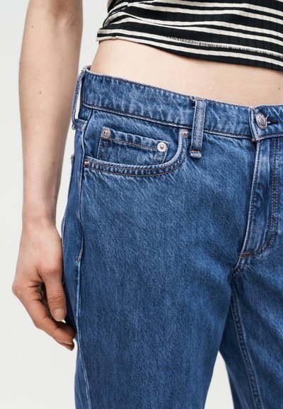 Άτομο με μπλε τζιν παντελόνι και μαύρο-λευκό ριγέ κομμένο τοπ, με το χέρι χαλαρό στο πλάι.