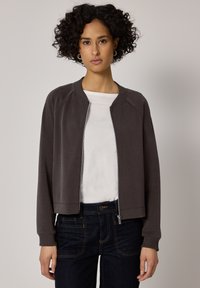 Frau mit kurzen lockigen Haaren, die goldene Creolen, eine dunkelgraue Reißverschlussjacke, ein weißes Hemd und dunkle blaue Jeans trägt, steht vor einem einfarbigen Hintergrund.