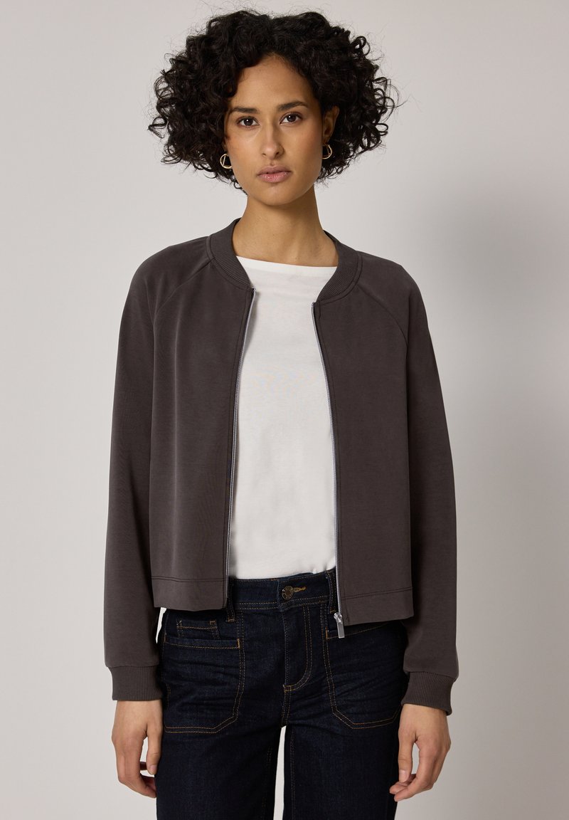 Frau mit kurzen lockigen Haaren, die goldene Creolen, eine dunkelgraue Reißverschlussjacke, ein weißes Hemd und dunkle blaue Jeans trägt, steht vor einem einfarbigen Hintergrund.