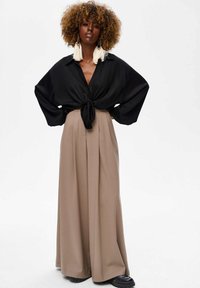 Zwarte blouse met lange mouwen en een geknoopte voorkant, gecombineerd met hoogwaist beige wijde broek en zwarte plateau schoenen.