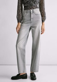 Svijetlosive visoke traperice s ravnim nogavicama. Istrošeni izgled, prednji džepovi i metalno kopčanje. Uparene s crnim cipelama.