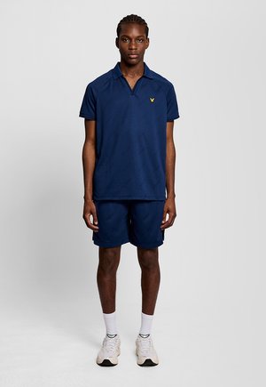 Ung man står i en marinblå pikétröja och matchande shorts, vita strumpor och vita sneakers mot en enfärgad grå bakgrund.