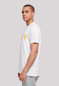 White cotton t-shirt with short sleeves, featuring a yellow rubber duck graphic on the left chest. Model is seen from the side.