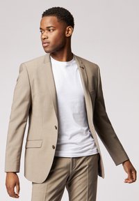 Hellbeige Anzugjacke mit strukturierten Schultern, zwei Knöpfen und Seitentaschen, getragen über einem weißen T-Shirt. Glatte Textur und taillierte Passform.