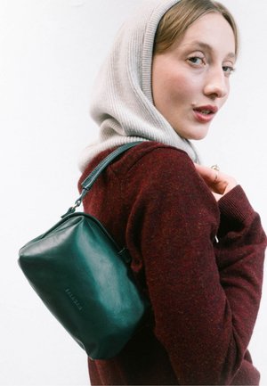 Sac en cuir vert en bandoulière de forme arrondie, avec fermeture éclair et bandoulière ajustable, porté avec un pull bordeaux à capuche grise claire.