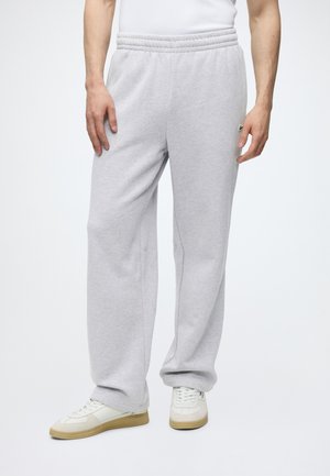 Uomo con pantaloni della tuta grigio chiaro e scarpe da ginnastica bianche, in piedi con le mani rilassate lungo i fianchi su sfondo bianco.