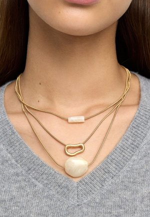 Naszyjniki warstwowe w złotym odcieniu o różnych fakturach. Jeden ma gładką owalną zawieszkę, inny prostokątny koralik, a kolejny rzeźbiony ogniwo.