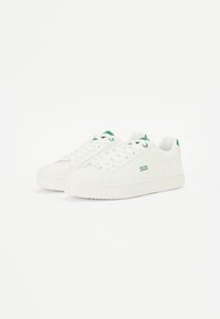 White canvas sneakers featuring green accents, round toe, flat sole, and textured rubber outsole. Logo printed on side.