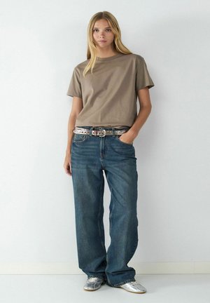Junge Frau mit langen blonden Haaren, die ein taupefarbenes T-Shirt, weite blaue Jeans, silbernen Gürtel und silberne Schuhe trägt und vor einer schlichten weißen Wand steht.