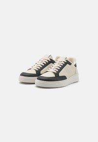 Baskets lacées avec un dessus en cuir beige, accents noirs, détails perforés et une semelle blanche rembourrée. Doublure texturée pour plus de confort.