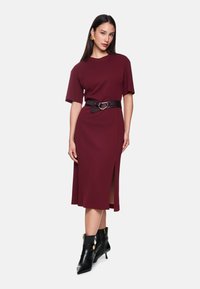 Bordeauxfarbenes Midikleid mit kurzen Ärmeln und seitlichem Schlitz, tailliert mit einem dunkelbraunen Gürtel mit einer runden Schnalle. Schwarze Knöchelstiefel.