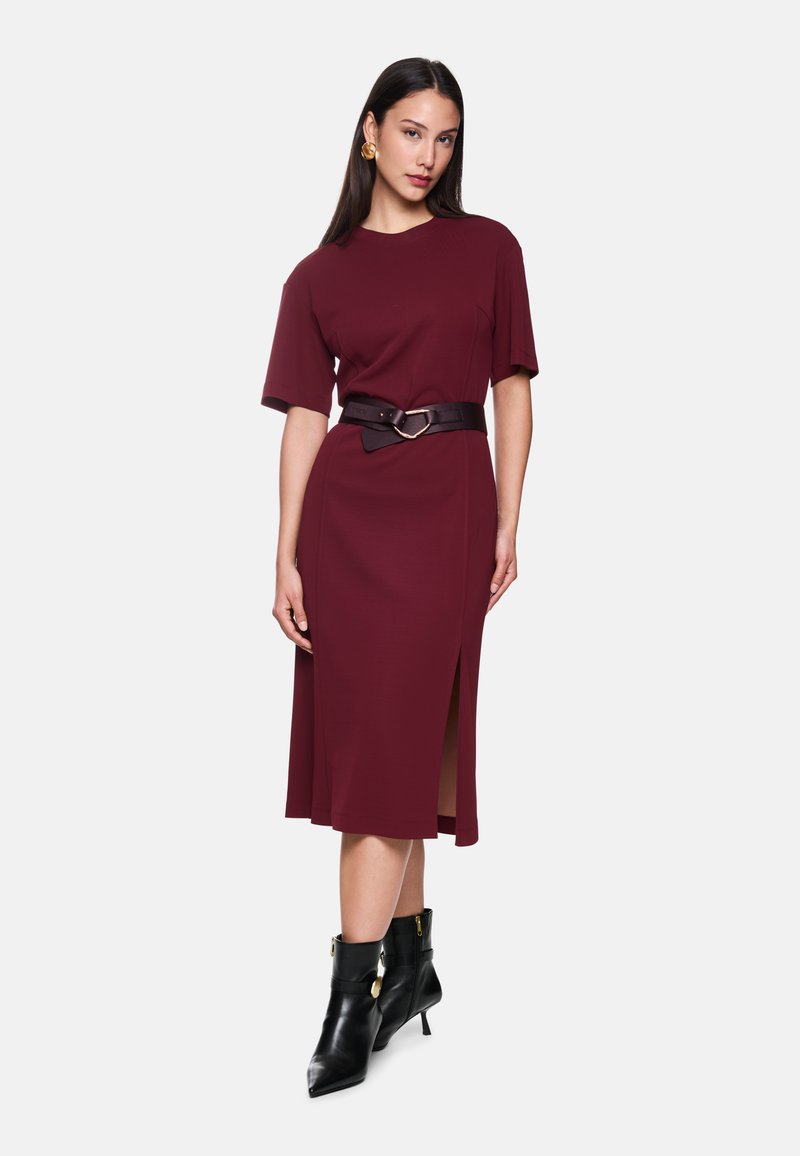 Bordeauxfarbenes Midikleid mit kurzen Ärmeln und seitlichem Schlitz, tailliert mit einem dunkelbraunen Gürtel mit einer runden Schnalle. Schwarze Knöchelstiefel.