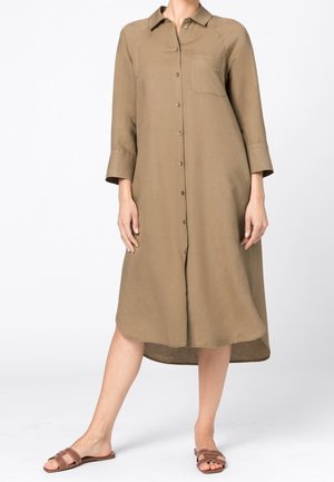 Kvinde iført en løs, langærmet, beige midi skjortekjole med knapper, brystlomme og brune flade sandaler.